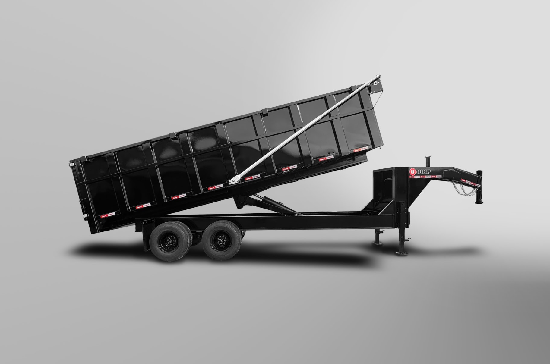 A black dump trailer with tandem wheels is shown on a plain gray background. The trailer bed is raised at an angle, indicating its dumping mechanism.