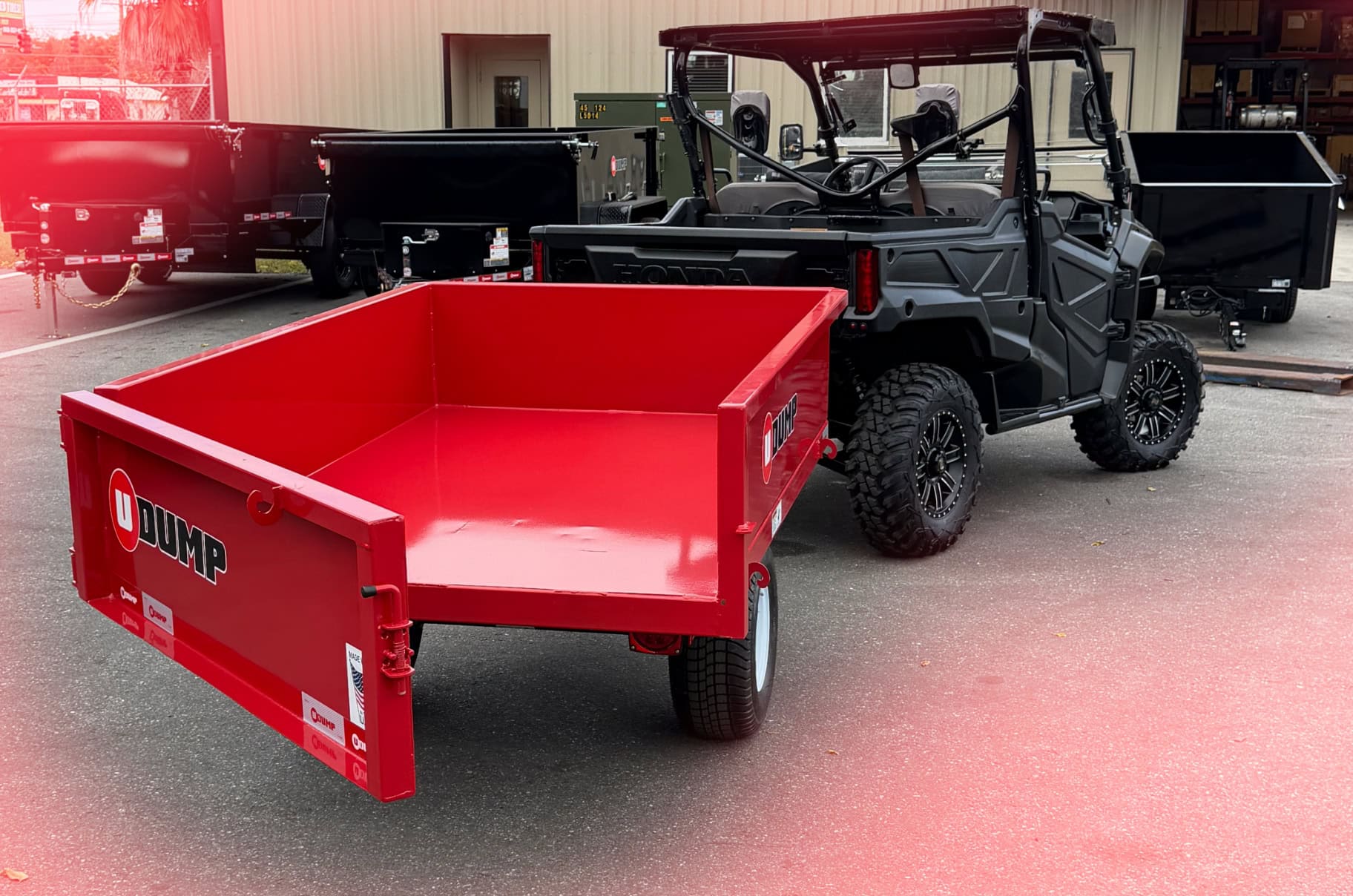 A black utility vehicle with a red dump trailer attached is parked on asphalt near other trailers and a tan industrial building.