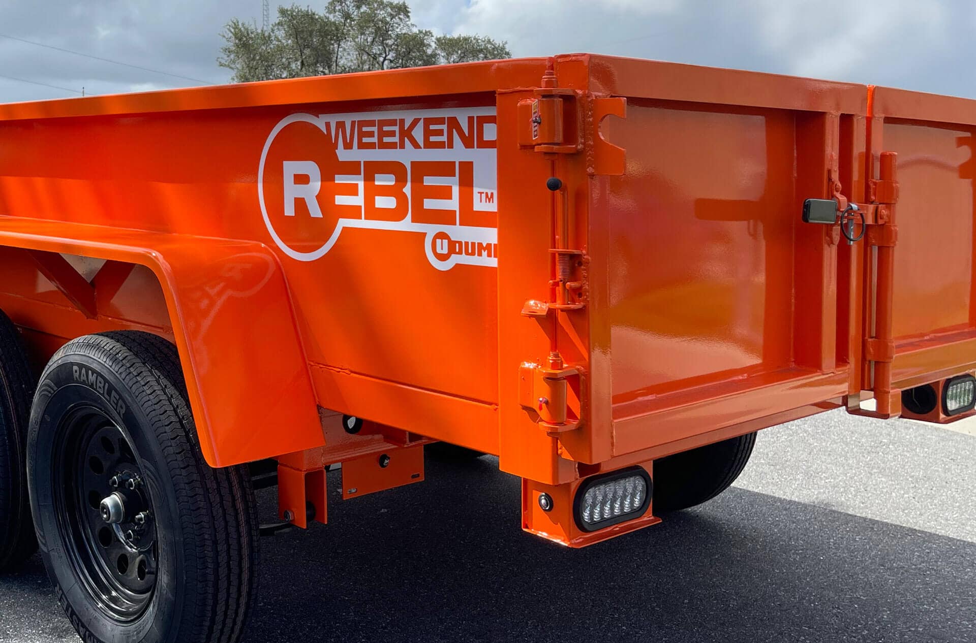 Close-up of a Rebel Mini Dump Trailer in orange with black wheels, featuring "WEEKEND REBEL" branding on the side, parked on pavement.