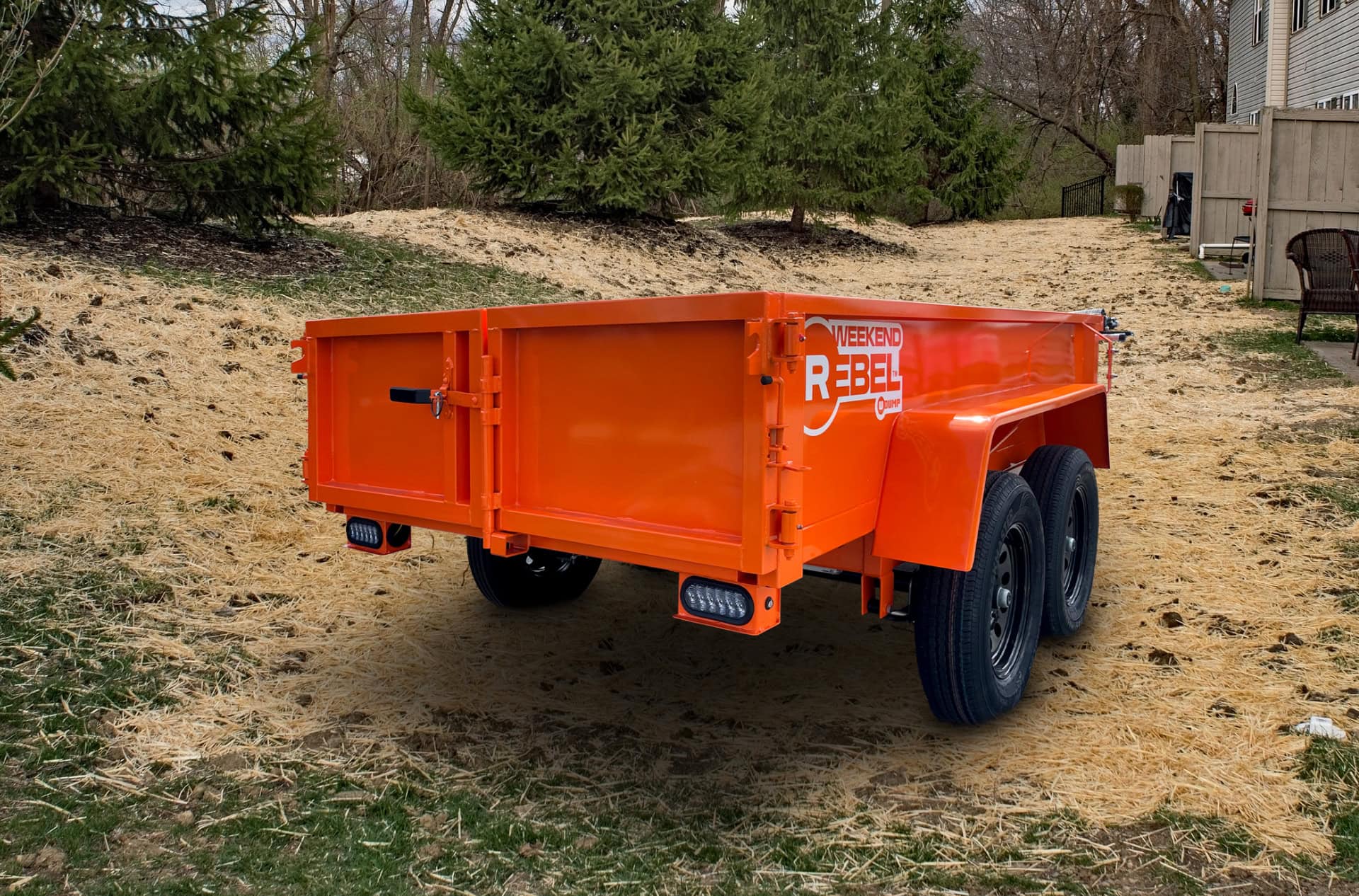 A bright orange Rebel Mini Dump Trailer with dual axles is parked on a grassy yard beside a house and trees; "Rebel" branding is visible on the side.